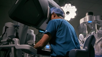 Surgeon operating a robotic surgical console, robotic arms and theatre lights behind
