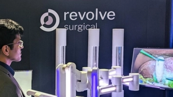 Person examining Revolve Surgical robotic system with illuminated arms beside a surgical procedure monitor